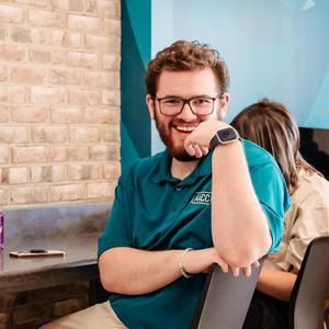 Gavin Kesselring sitting at a table smiling. He's wearing a teal AACC polo.